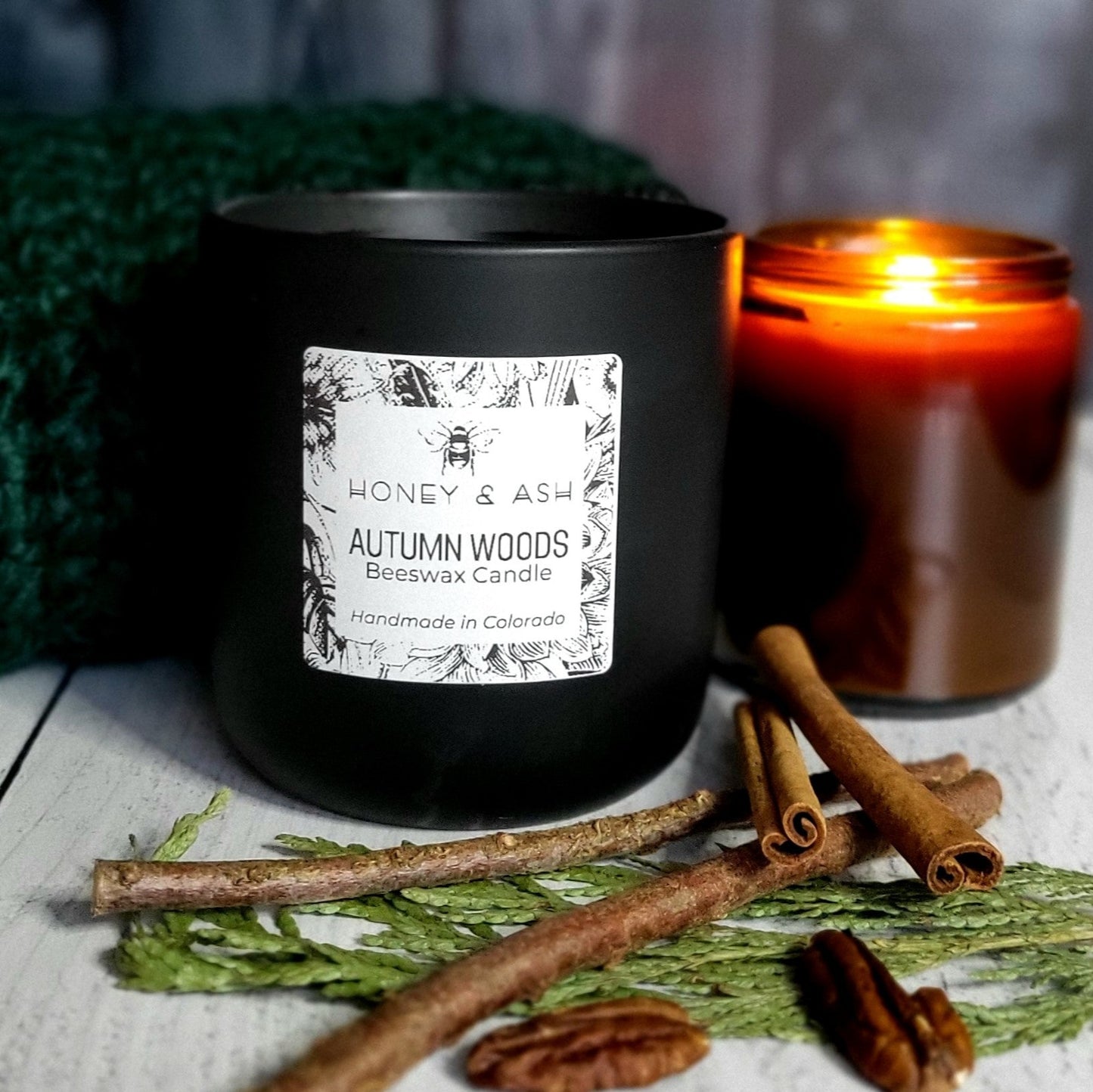 Front view of an Autumn Woods candle in a 12ox Black Ceramic Tumbler, in front of a green knitted blanket and lighted amber jar candle, with its scent ingredients in the foreground.