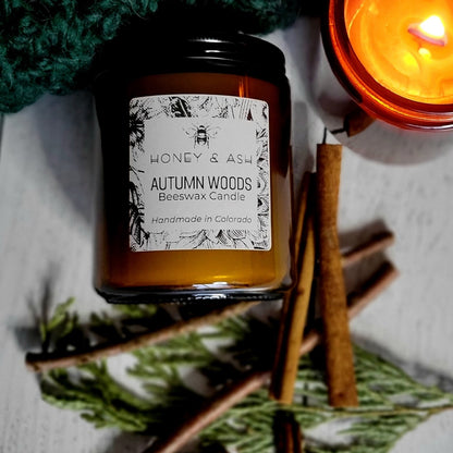 Side view of 8oz Amber Jar candle scented Autumn Woods, surrounded with its scent ingredients, leaning against a green knitted blanket, and next to a lighted candle,.